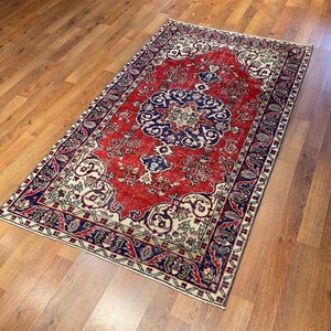 Pu&ograve; includere: Un tappeto rettangolare caratterizzato da un campo centrale rosso vibrante con un prominente medaglione circolare blu scuro. Il tappeto &egrave; decorato con intricati motivi floreali e a viticcio in crema, blu navy e azzurro, incorniciato da un'ampia bordura blu scuro.