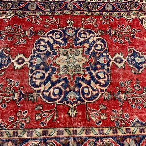 Pu&ograve; includere: Un tappeto rettangolare caratterizzato da un campo rosso vivace con un medaglione centrale blu scuro. Il design include complessi motivi floreali e geometrici in tonalit&agrave; crema, azzurro e blu scuro, rifinito con un bordo dettagliato.