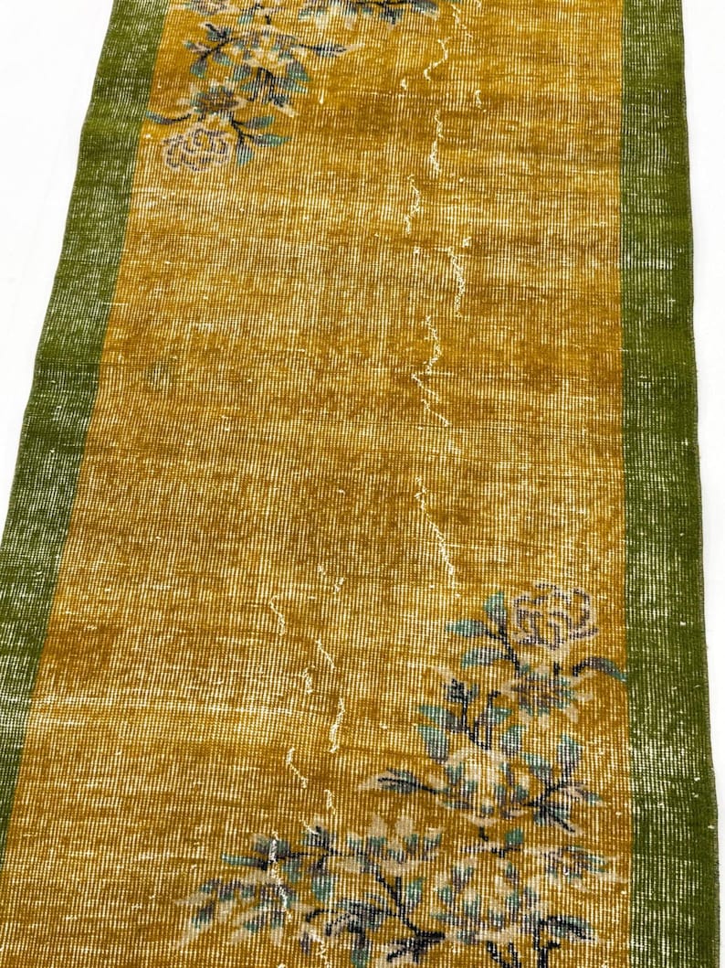 May include: A rectangular runner rug featuring a mustard yellow centre field with a distressed texture and faded floral motifs in blue and white. The rug is framed by solid green borders on both long sides, showcasing a worn, vintage appearance.