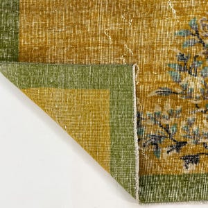 May include: A rectangular area rug featuring a mustard yellow field with a green border. The rug displays a floral pattern in shades of light blue, white, and dark grey. One corner of the rug is folded over, revealing the underside of the fabric.