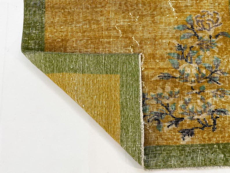 May include: A rectangular area rug featuring a mustard yellow field with a green border. The rug displays a floral pattern in shades of light blue, white, and dark grey. One corner of the rug is folded over, revealing the underside of the fabric.