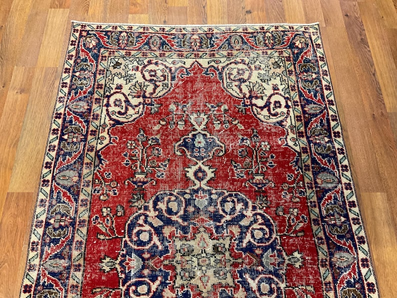 Pu&ograve; includere: Un tappeto rettangolare caratterizzato da un campo centrale rosso vivace con intricati motivi floreali blu e crema. Il tappeto &egrave; incorniciato da un bordo ampio e dettagliato nei toni del blu profondo, rosso e crema, con motivi simmetrici.