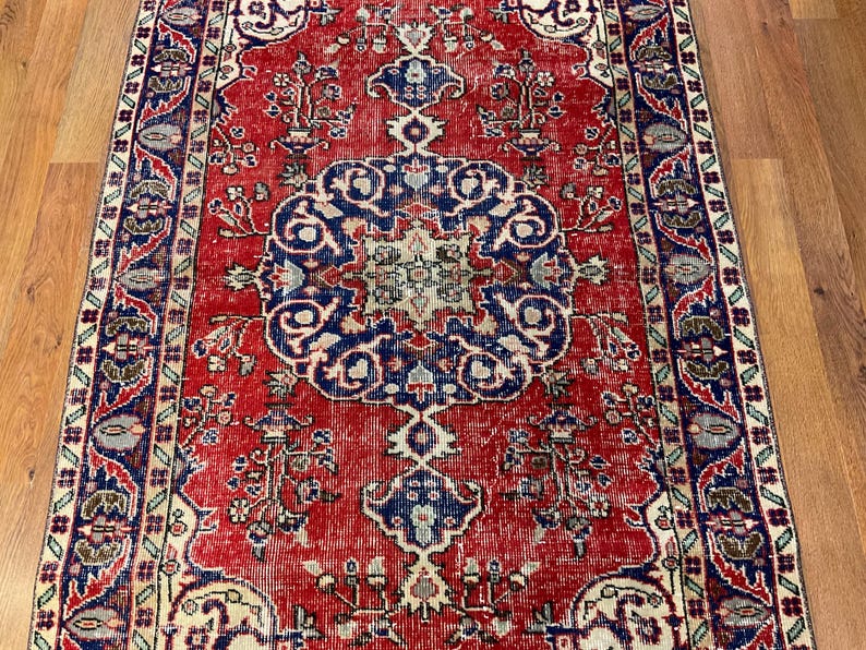 Pu&ograve; includere: Un tappeto rettangolare caratterizzato da un campo rosso vivace con un grande medaglione centrale blu scuro. Il design presenta intricati motivi floreali, schemi simmetrici e un bordo decorativo nei toni del blu, crema e verde spento.