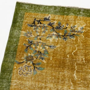 May include: A rectangular rug featuring a mustard yellow field with a green border. The rug displays a floral pattern in shades of teal, black, and white, with a visible woven texture and a frayed edge along the top border of the fabric.