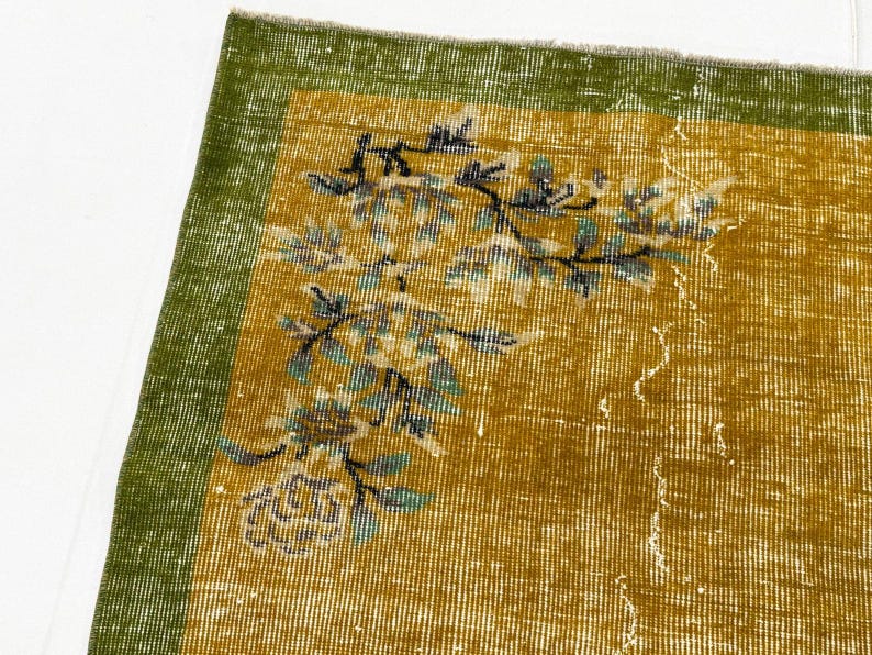 May include: A rectangular rug featuring a mustard yellow field with a green border. The rug displays a floral pattern in shades of teal, black, and white, with a visible woven texture and a frayed edge along the top border of the fabric.