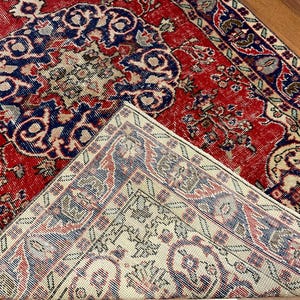 Pu&ograve; includere: Un tappeto rettangolare con uno sfondo rosso vivace e intricati motivi floreali e geometrici in blu, crema e marrone scuro. Un angolo del tappeto &egrave; ripiegato, rivelando un retro color crema pi&ugrave; chiaro con un motivo corrispondente.