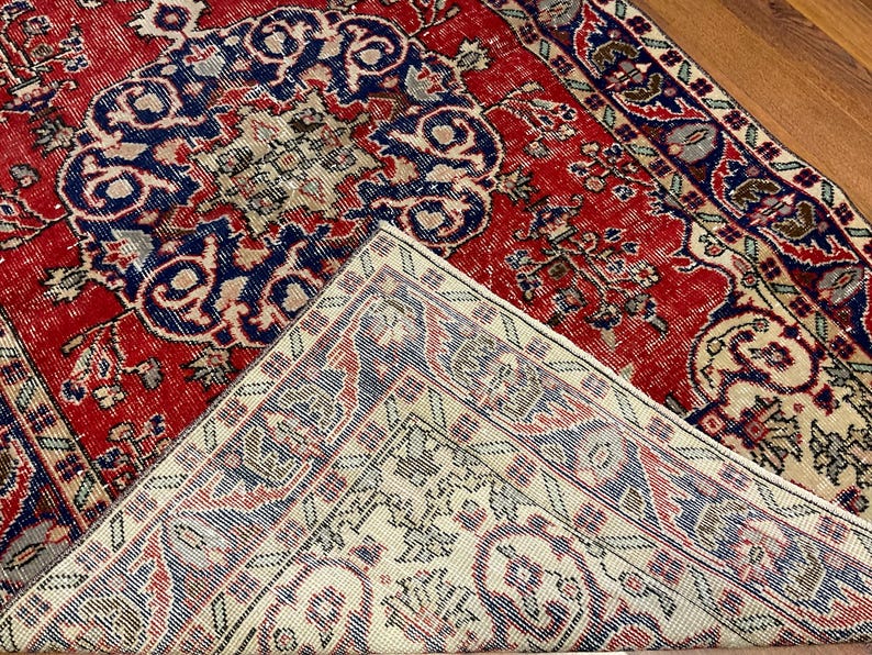 Pu&ograve; includere: Un tappeto rettangolare con uno sfondo rosso vivace e intricati motivi floreali e geometrici in blu, crema e marrone scuro. Un angolo del tappeto &egrave; ripiegato, rivelando un retro color crema pi&ugrave; chiaro con un motivo corrispondente.