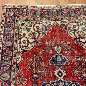 Pu&ograve; includere: Un tappeto rettangolare caratterizzato da un campo centrale rosso intenso con intricati motivi floreali e geometrici in blu navy, crema e verde salvia. Il bordo presenta un motivo ripetitivo di fiori e viticci stilizzati in una palette multicolore.