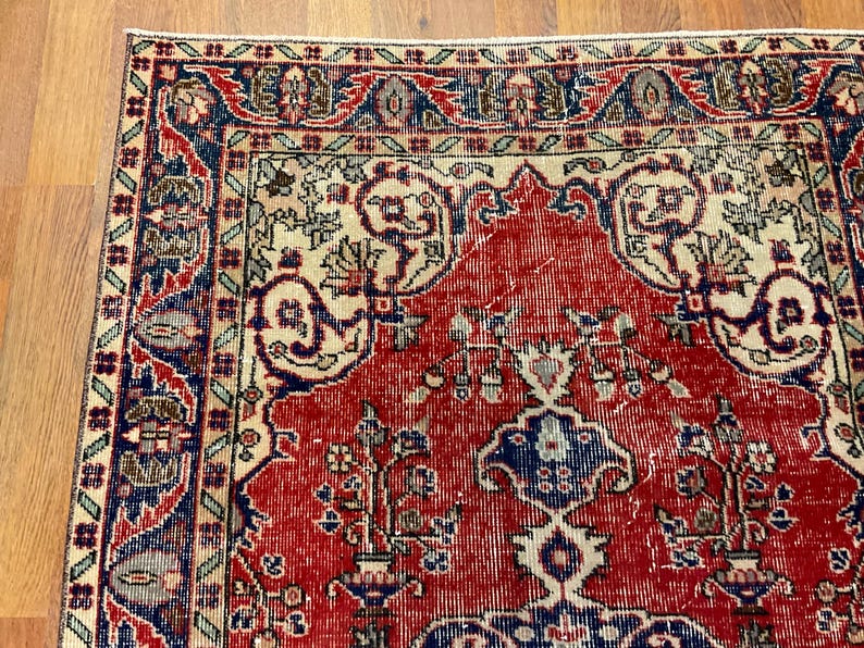 Pu&ograve; includere: Un tappeto rettangolare caratterizzato da un campo centrale rosso intenso con intricati motivi floreali e geometrici in blu navy, crema e verde salvia. Il bordo presenta un motivo ripetitivo di fiori e viticci stilizzati in una palette multicolore.