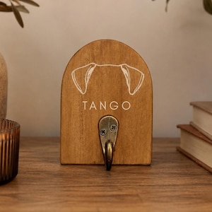 May include: An arched wooden wall hook featuring a natural brown finish. The front displays a white line drawing of dog ears and the name TANGO printed in white capital letters. A single antique brass-colored metal hook is attached at the bottom.