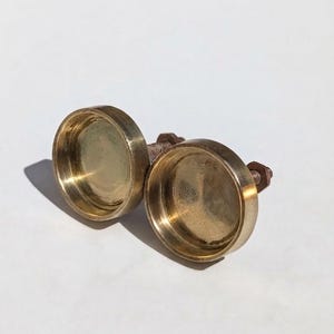 May include: Two circular brass knobs with a recessed center and a threaded metal bolt extending from the back. The knobs have a polished metallic finish with a warm golden hue and a smooth, rounded edge profile for attachment to furniture.