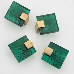 Può includere: Quattro pomelli quadrati per armadietti con un pannello frontale in vetro smeraldo traslucido e testurizzato, abbinato a una base in metallo solido color ottone spazzolato. Ogni pomello presenta un motivo superficiale irregolare e un attacco quadrato.