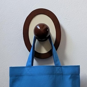 Può includere: Un gancio da parete in legno con un pomello marrone scuro e un design circolare. Una borsa blu è appesa al gancio. Il gancio è montato su un muro bianco.