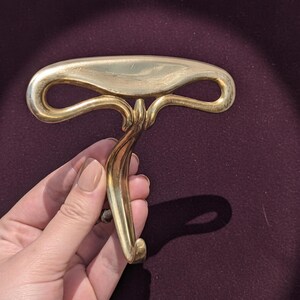 May include: A gold-colored, ornate handle with a curved design. The handle has a central stem that curves into a hook shape. The handle is held against a dark purple background.