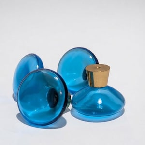 May include: A set of four round, translucent blue glass cabinet knobs, each featuring a smooth, circular face and a metallic gold-toned cylindrical base. The knobs are arranged in a cluster, showcasing their vibrant blue color and reflective surface.