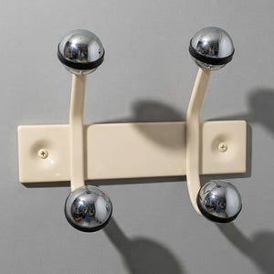 以下が含まれることがあります： クリーム色の長方形のベースに2本の取り付けネジが付いた壁掛け式コートラック。ベースから2本の曲線的なアームが伸びており、それぞれに黒いゴムリングが付いた光沢のあるクロームメッキの球体フックが2つずつ取り付けられています。