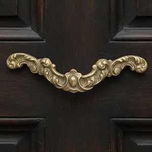 Könnte beinhalten: Ein messingfarbener Schubladengriff aus Metall mit einem verzierten, geschwungenen Design, das symmetrische Schnörkel und ein zentrales ovales Detail aufweist. Der Beschlag ist horizontal auf einer dunkelbraunen Holzoberfläche montiert.