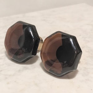 May include: Two hexagonal cabinet knobs made of translucent dark brown glass. Each knob features a faceted surface with a smooth, polished finish and a central circular indentation. The knobs are mounted on a small, metallic base component.