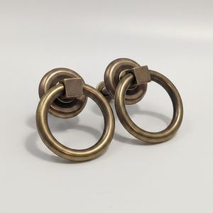 Puede incluir: Dos tiradores de anillo de latón antiguo para muebles. Cada tirador presenta un anillo circular unido a una base cuadrada y una placa trasera redonda. La ferretería tiene un acabado cálido y envejecido, lo que sugiere una estética vintage o rústica.