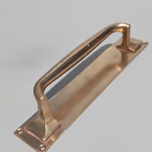 May include: A polished brass door pull handle featuring a long, rectangular mounting plate with two visible screw holes at each end. The handle has a smooth, arched design that connects to the base plate at two points, creating a sturdy grip.