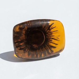 Puede incluir: Un anillo rectangular de resina color ámbar con un patrón radial oscuro en forma de rayos de sol que emana de una hendidura circular central. La pieza tiene un acabado liso, pulido y translúcido con tonos de color marrón dorado.