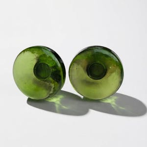 May include: Two circular, translucent olive green glass candle holders with a smooth, polished surface. Each piece features a central cylindrical indentation designed to securely hold a standard taper candle in an upright position.