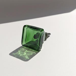 May include: A square, green glass bottle stopper with a dark metal base. The stopper is transparent, allowing light to pass through and cast a patterned shadow. The product is displayed on a white surface.
