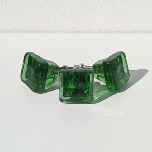 Peut inclure: Trois boutons de meuble carrés en verre vert émeraude avec des fixations argentées. Les boutons sont disposés sur une surface blanche, projetant des ombres. Ces boutons décoratifs sont parfaits pour ajouter une touche de couleur aux meubles.
