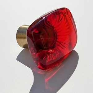 Puede incluir: Un tirador de armario cuadrado de vidrio rojo con base de latón. El tirador es de un rojo vibrante y transparente, y la base de latón es de color oro cálido. El tirador se utiliza probablemente para muebles o armarios.