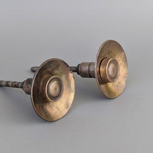 May include: Two antique brass cabinet knobs with a weathered finish. Each knob features a circular, flat plate with a raised centre and a threaded post for mounting. The knobs have a vintage, aged appearance.