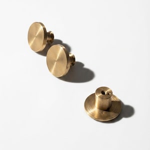Op de afbeelding: Drie ronde kastknoppen van messing met een geborstelde metalen afwerking. Elke knop heeft een plat, rond oppervlak en een cilindrische basis met een schroefgat in het midden voor montage. De knoppen zijn in een diagonale lijn geplaatst.