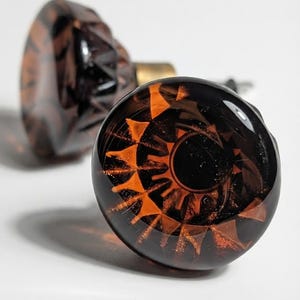 May include: Two round cabinet knobs made of dark brown, translucent glass. Each knob features a faceted, star-like pattern embedded within the glass, creating a deep amber and black contrast. A metallic base is visible on the rear of each knob.