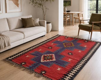 Op de afbeelding: Een rechthoekig rood vloerkleed met een geometrisch patroon in blauw, zwart en beige. Het vloerkleed heeft een gefranjerde rand en ligt op een houten vloer in een woonkamer. Het ontwerp van het vloerkleed bevat ruit- en geometrische vormen.