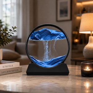 May include: A circular sand art display featuring a black frame and base. Inside the glass, blue and white sand forms a mountain-like landscape with cascading sand particles creating a waterfall effect against a clear liquid background.