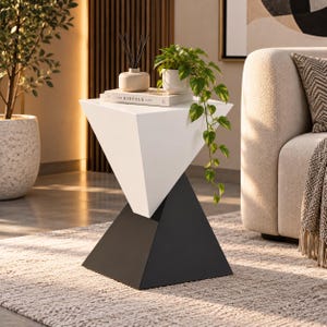 Puede incluir: Una mesa auxiliar con una parte superior triangular blanca equilibrada sobre una base triangular negra. Sobre la mesa hay un libro blanco titulado The Kinfolk Home, un pequeño difusor beige con varillas negras y una maceta blanca con una planta.