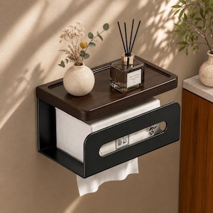 Puede incluir: Un dispensador de pañuelos de pared con marco de metal negro mate y estante superior de madera marrón oscuro. La unidad sostiene una caja rectangular de pañuelos blancos, con una ranura horizontal. Un pequeño jarrón beige y un difusor están encima.