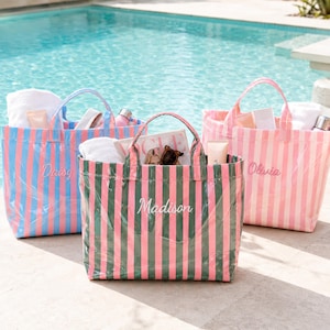Peut inclure: Trois sacs fourre-tout rectangulaires avec deux anses, ornés de rayures verticales. Le sac de gauche est bleu et rose avec le nom Daisy, le sac central est vert et rose avec le nom Madison, et le sac de droite est rose et blanc avec le nom Olivia.