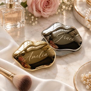 May include: Two lip-shaped trinket dishes with metallic finishes. One dish is gold-toned with the name Amelia engraved in script, and the other is silver-toned with the name Grace engraved in script. Both dishes feature smooth, reflective surfaces.