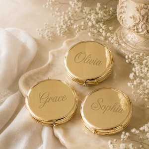 May include: Three round, polished gold-tone metal compact mirrors. Each mirror features a smooth, reflective surface with a name engraved in a cursive script font. The names engraved on the individual compacts are Olivia, Grace, and Sophia.