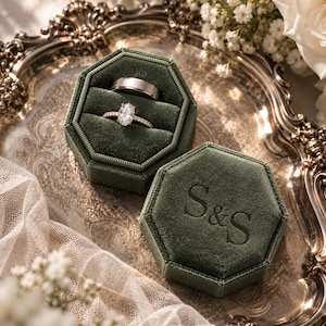 May include: An octagonal dark green velvet ring box with the letters S&S embossed on the lid. The open box contains two rings: one plain metallic band and one ring featuring a central oval-cut stone set on a thin, pavé-style band.