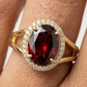 Può includere: Un anello in oro con una grande pietra preziosa granato rosso scuro a taglio ovale al centro. Il granato è circondato da un alone di piccoli diamanti bianchi rotondi e chiari. L'anello è realizzato in oro giallo lucido con un design a gambo diviso.