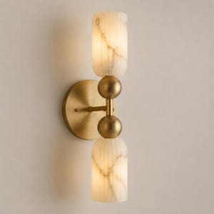 May include: A wall sconce featuring a circular brass backplate and a central vertical arm with two spherical brass accents. Two cylindrical, vertically ribbed, white marble-patterned glass shades are positioned at the top and bottom ends.