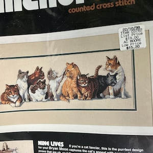 May include: A Dimensions counted cross stitch kit titled Nine Lives featuring a horizontal illustration of nine cats in various colors and poses on tan fabric. The packaging includes text, a color accented chart, and dimensions of 18 inches by 7 inches.