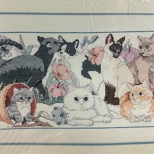 May include: A cross-stitch embroidery pattern featuring seven cats of various breeds and colors, including a gray tabby in a terracotta pot, a black cat, a tuxedo cat, a Siamese, a white long-haired cat, and an orange tabby, surrounded by pink flowers.