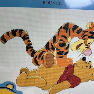 May include: A cross-stitch pattern featuring Tigger, an orange and black striped tiger, bouncing on top of Winnie the Pooh, a yellow bear wearing a red shirt. The word BOUNCE is printed in white at the top center against a light blue background.