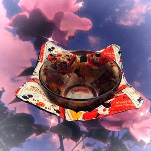May include: A fabric bowl cosy featuring a vintage Mickey Mouse comic strip print in red, black, and cream, holding a clear glass bowl. The cosy has four pointed corners and is constructed with quilted fabric panels displaying various Mickey Mouse illustrations.