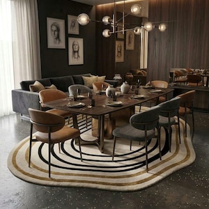 Puede incluir: Una alfombra ovalada con anillos concéntricos en crema, negro y oro metálico. La alfombra está colocada bajo una mesa de comedor rectangular de madera con sillas de tela beige y gris, sobre un suelo oscuro en un comedor.