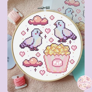Peut inclure: Broderie au point de croix dans un cercle en bois, représentant des images pixélisées de deux pigeons, du pop-corn, des nuages et des cœurs roses, violets et jaunes. Le motif est sur tissu blanc, avec des bobines de fil rose en arrière-plan.
