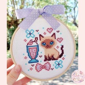 Peut inclure: Un cercle de broderie au point de croix avec un chat siamois, un milk-shake et des motifs floraux et en forme de cœur. Le design utilise du marron, du bleu, du rose et du violet, avec un ruban à pois violets clairs. Le cercle est tenu dans une main.