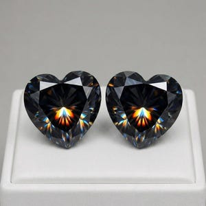 Può includere: Due gemme sfaccettate a forma di cuore, di colore grigio scuro. Le gemme sono esposte su una superficie bianca, su sfondo grigio. Le sfaccettature riflettono un arcobaleno di colori.